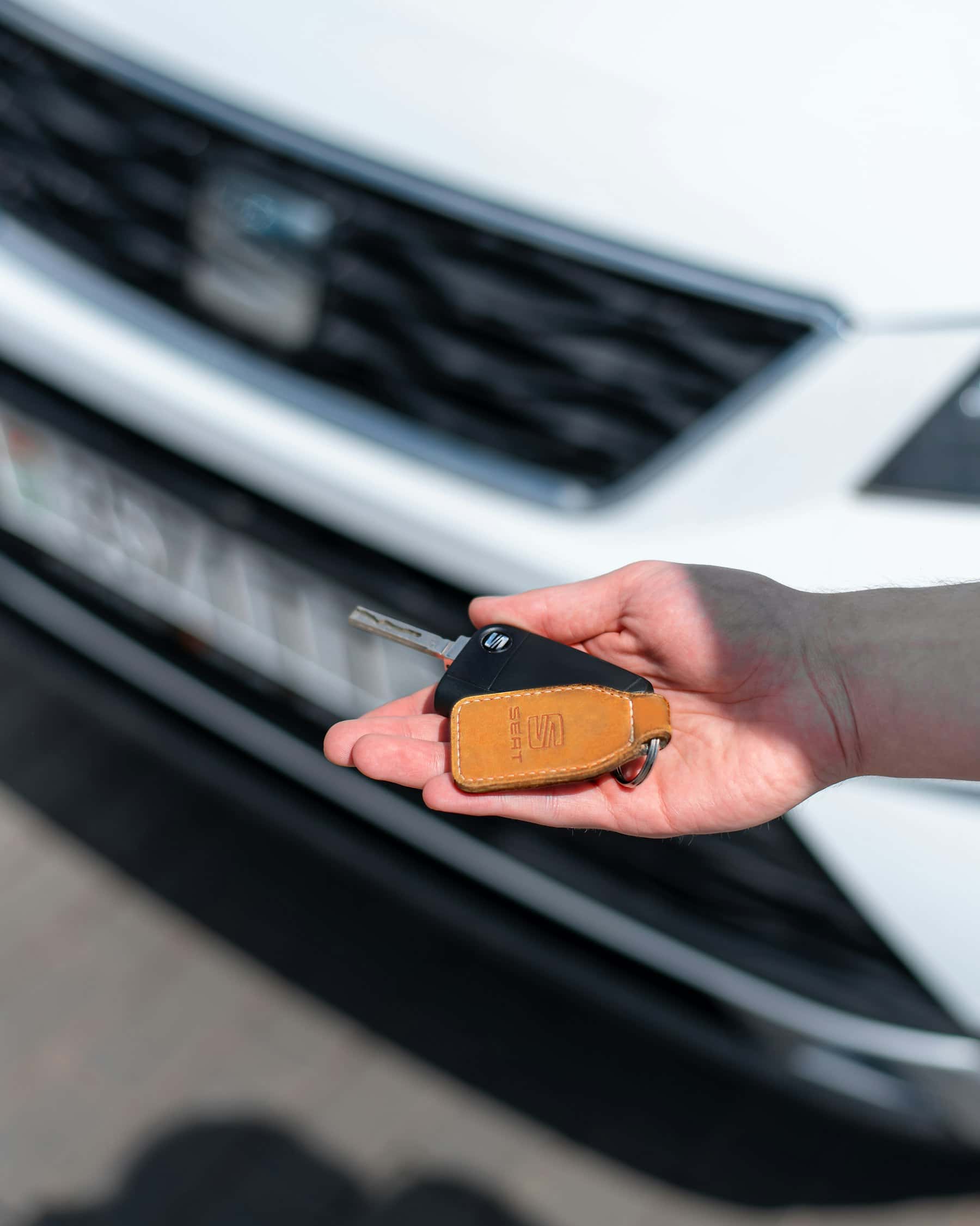 A hand holding out a car key and leather fob in front of a white car